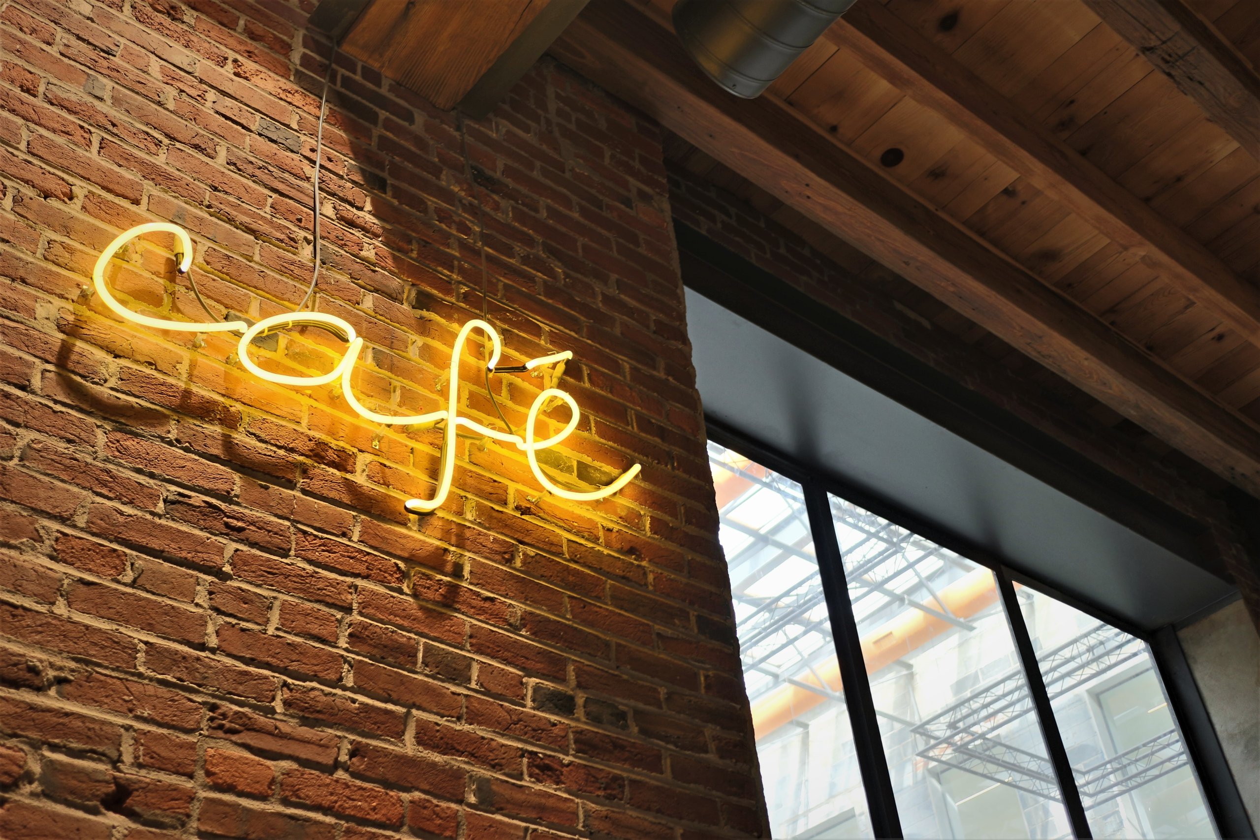 A neon script café sign on the original brick at Billy Penn Studios, with the atrium truss visible through the window