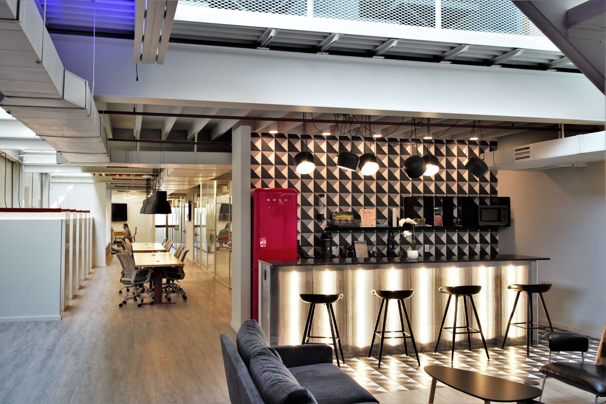 The coworking kitchen at Billy Penn Studios, a lit bar with stools and a geometric tiled backsplash, under the mezzanine and beside the glass conference room