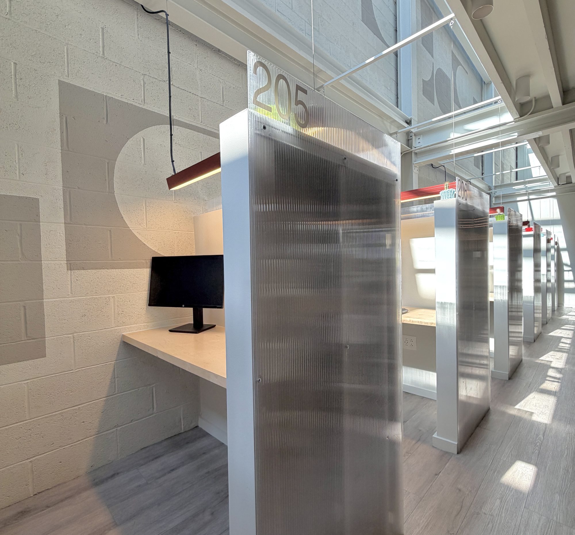A row of numbered dedicated desks at Billy Penn Studios coworking, each with an aluminium privacy screen, monitor and pendant lamp under a clerestory window