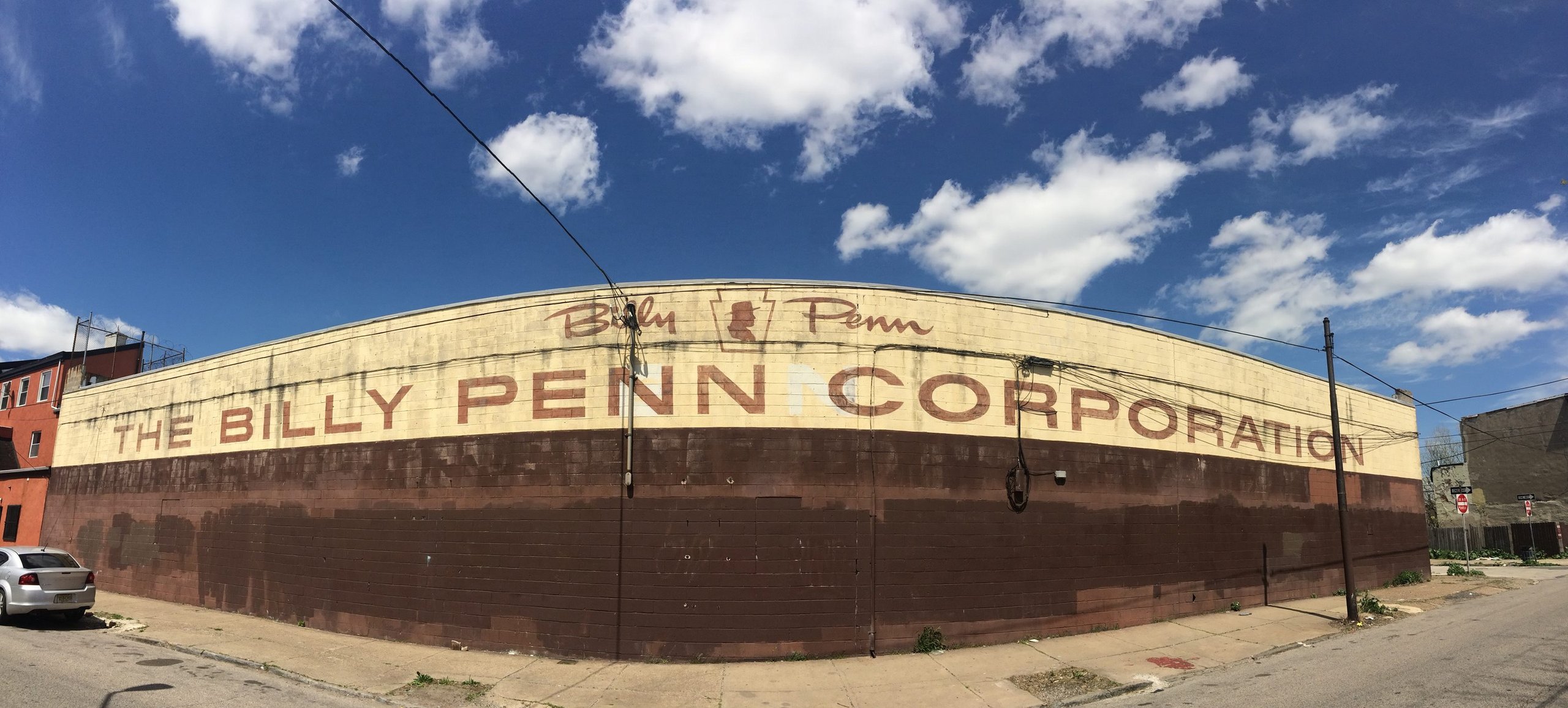 A panorama of the painted Billy Penn Corporation sign wrapping the corner of the building in Olde Kensington, Philadelphia